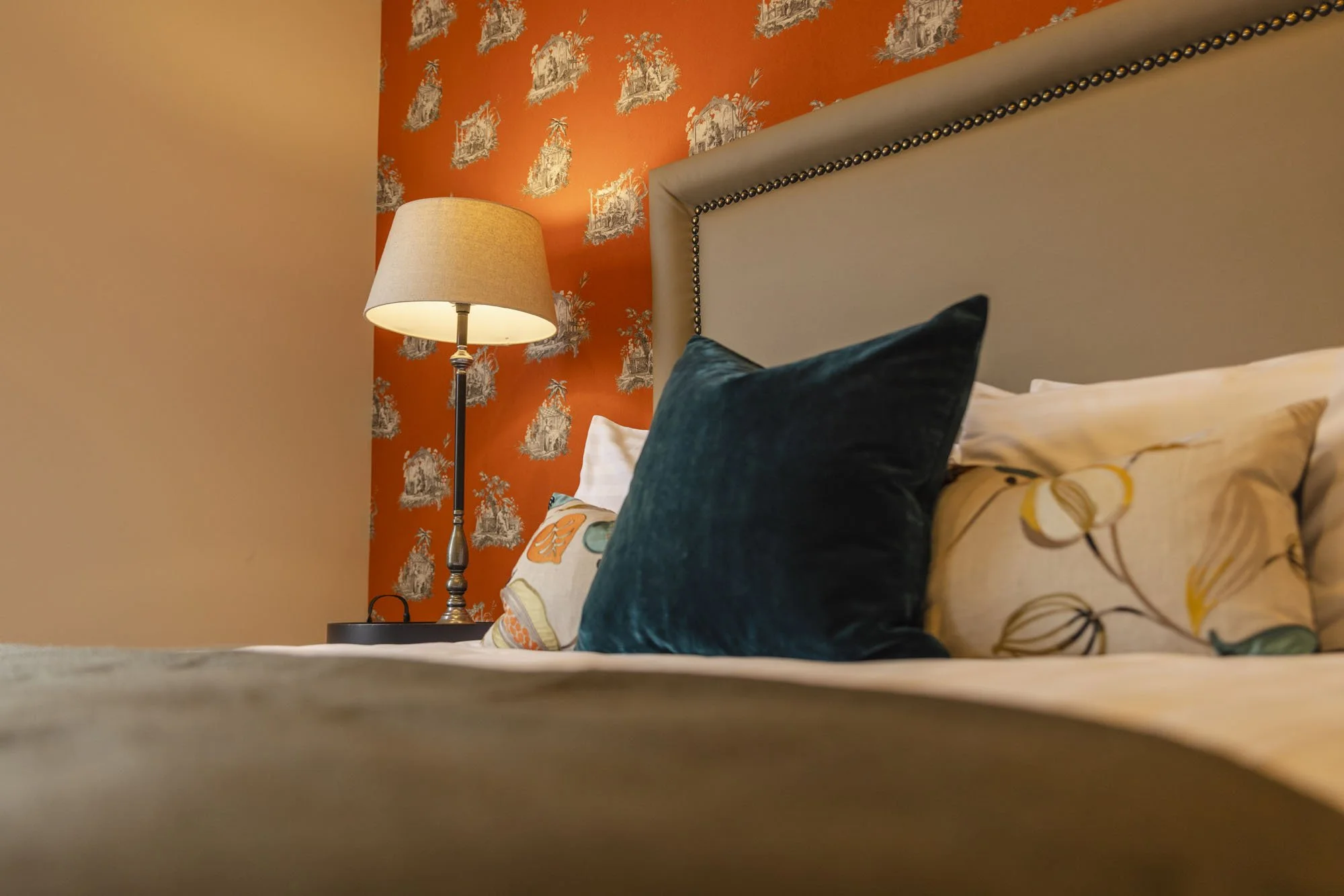 Close-up of a bedroom with orange wallpaper featuring white pastoral scenes, a beige headboard with nailhead trim, various colorful pillows, and a bedside lamp with a beige shade.