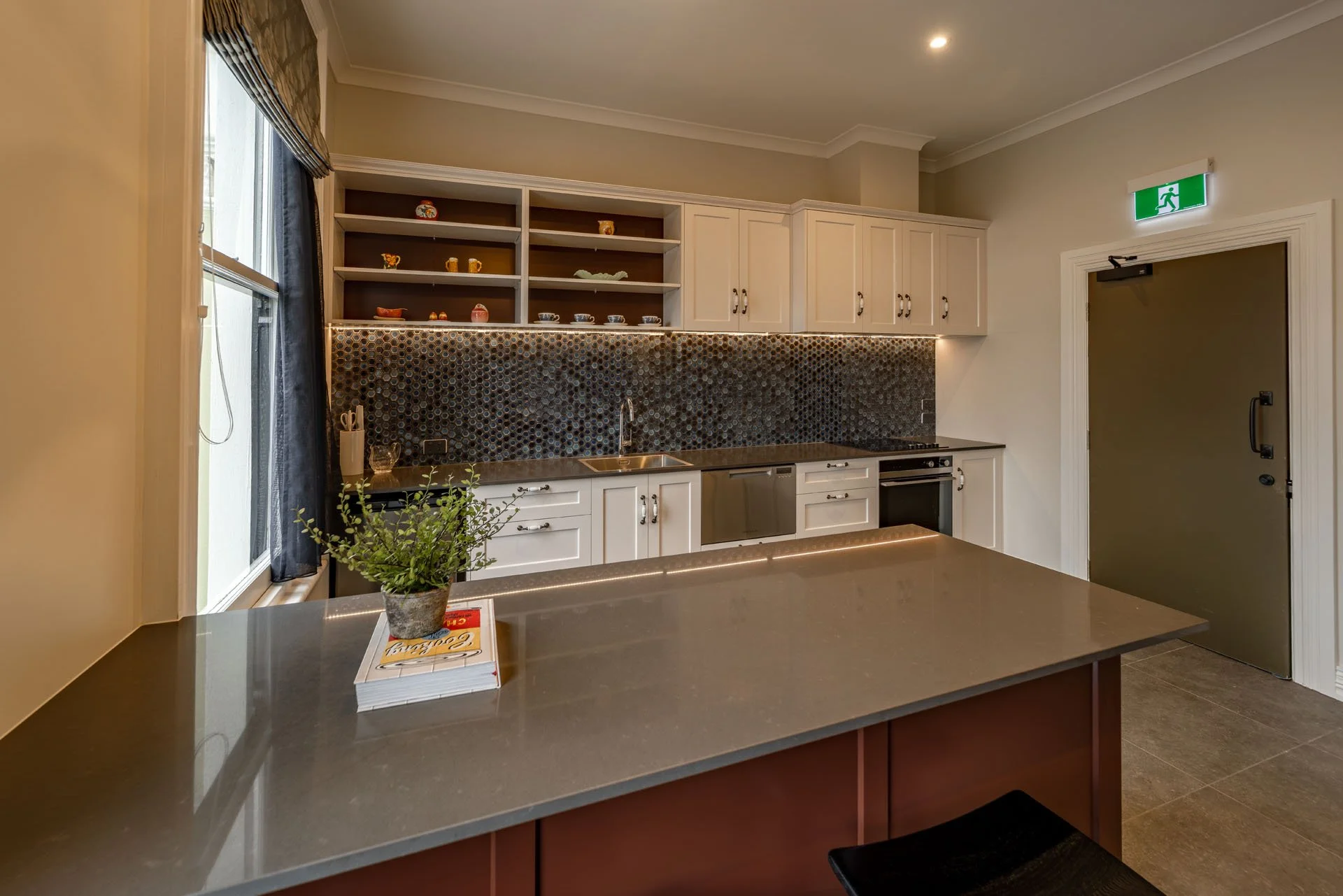 Modern kitchen with white cabinets, black backsplash, open shelving with dishes, a sink, and a kitchen island with a potted plant on top.