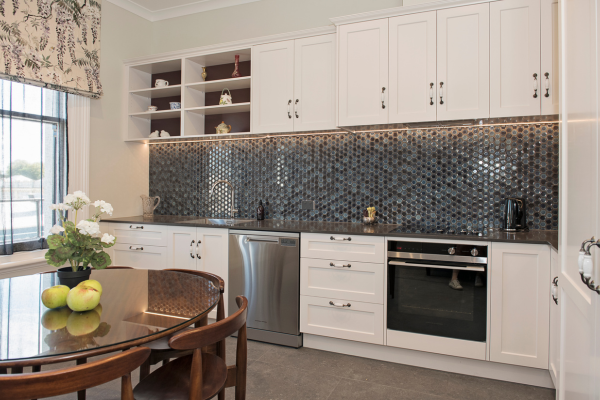 Kitchen with white cabinets, dark mosaic tile backsplash, stainless steel dishwasher, oven, black kettle, and a round wooden dining table with chairs, decorated with apples and flowers.