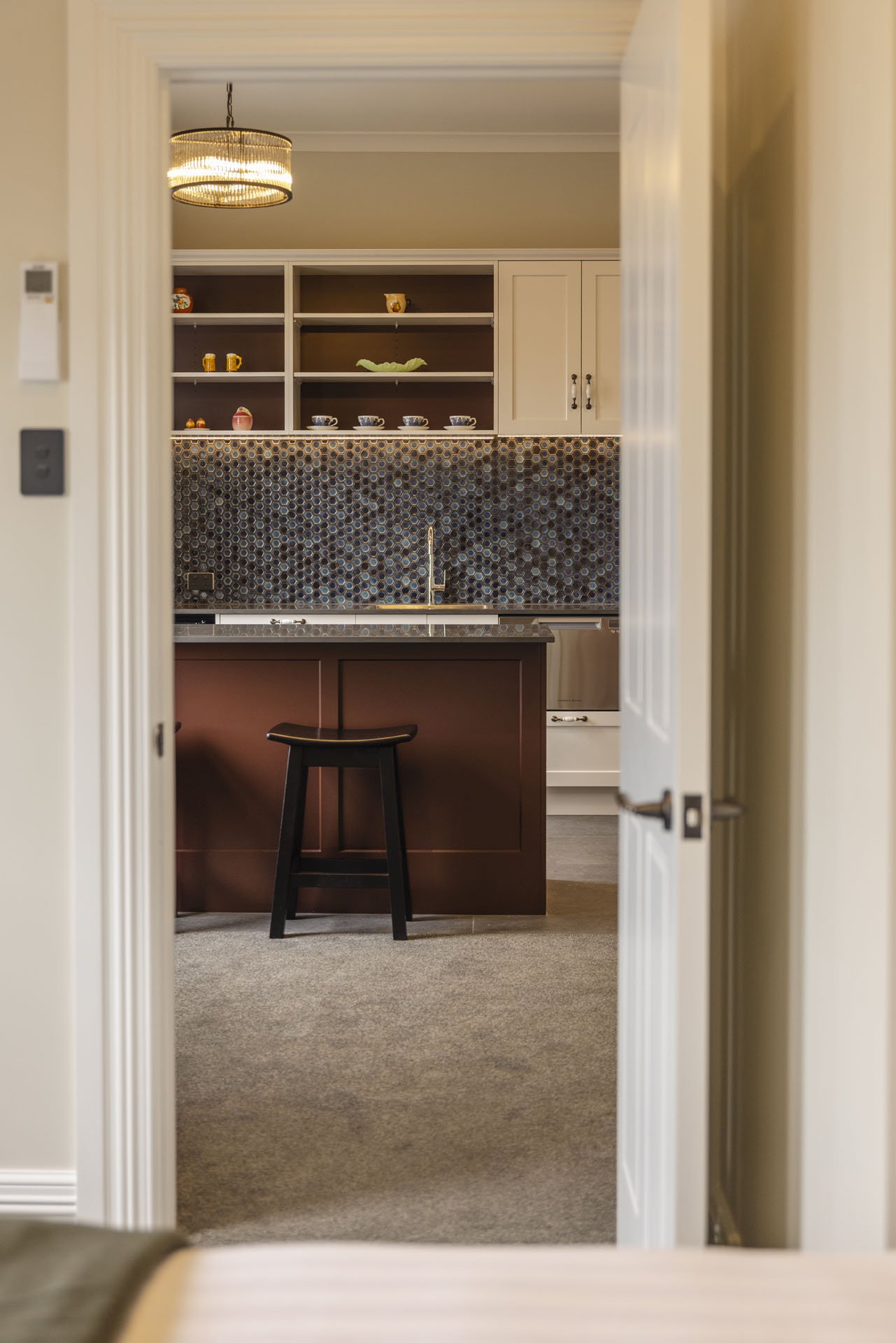 View of a kitchen seen through an open doorway, featuring a dark island with a black stool, an mosaic tile backsplash, and open shelves with decorative items and dishes.