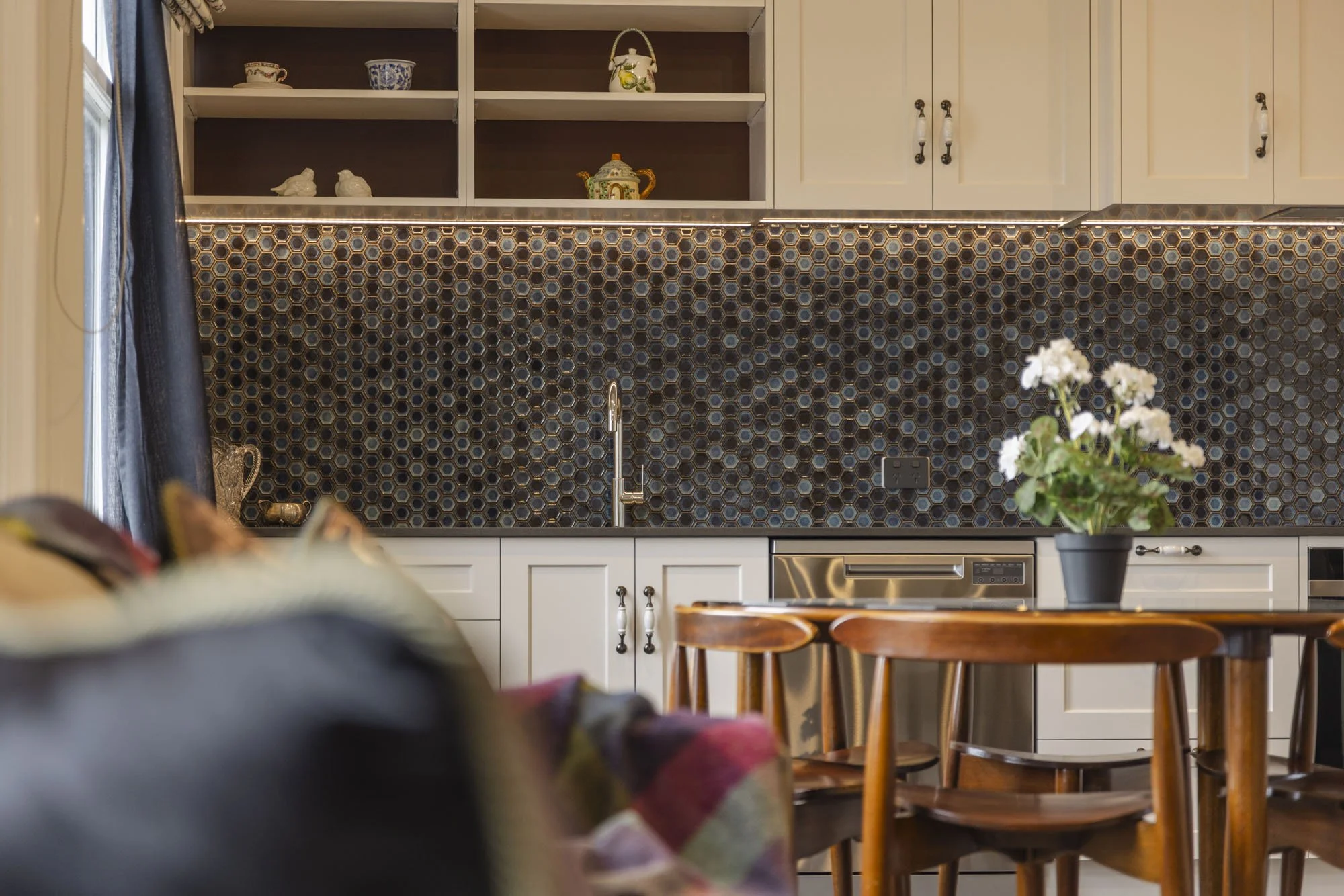 A kitchen with dark honeycomb tile backsplash, white cabinets, a stainless steel dishwasher, and a wooden dining table with matching chairs. A potted white flower on the table and open shelves with decorative items are also visible.