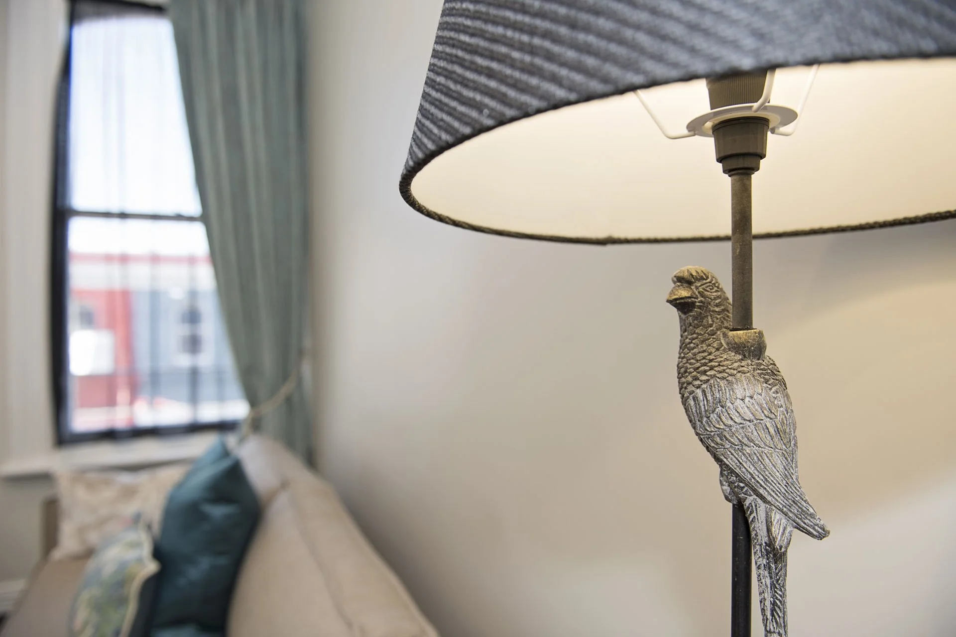 Close-up of a decorative table lamp featuring a carved bird figure as the base, with a beige lampshade, in a living room with a sofa and window with curtains in the background.