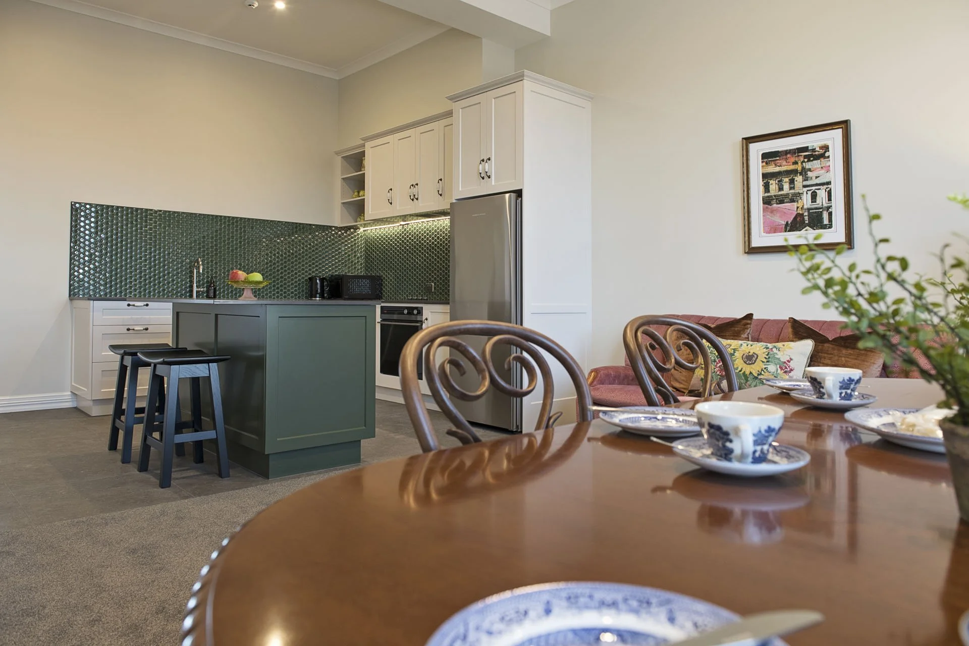 View of a modern kitchen and dining area featuring a round wooden dining table set with blue and white patterned dishes, a pink sofa with pillows, a framed artwork on the wall, white upper cabinets and green lower cabinets in the kitchen, a green til