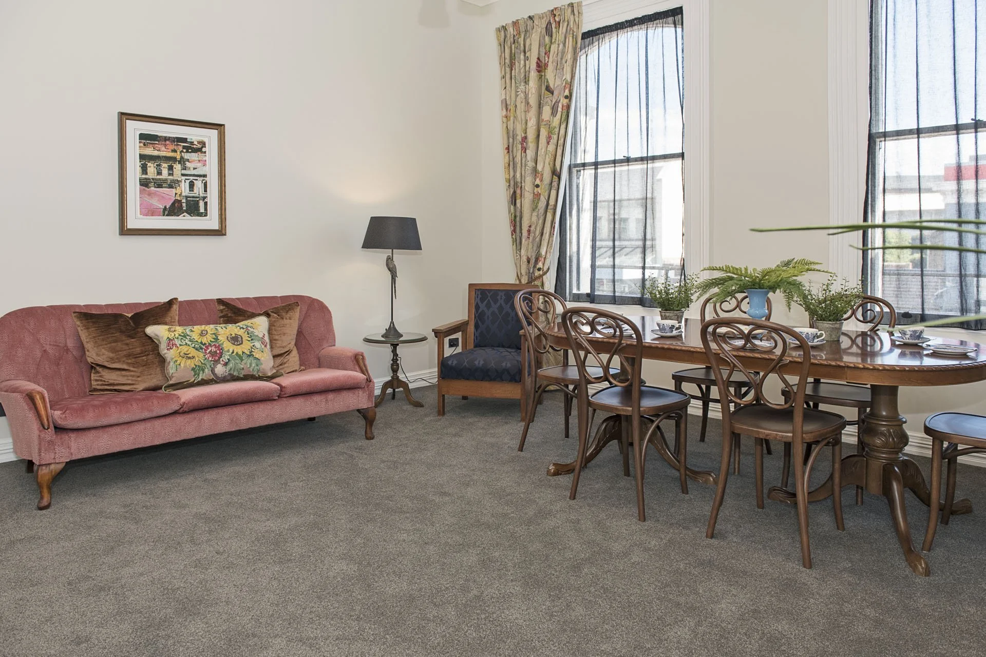 Living room with pink vintage sofa adorned with floral and brown cushions, a small black table lamp on a stand, a framed picture on the wall, a wooden dining table with six chairs, three potted plants, and large windows with floral curtains and black