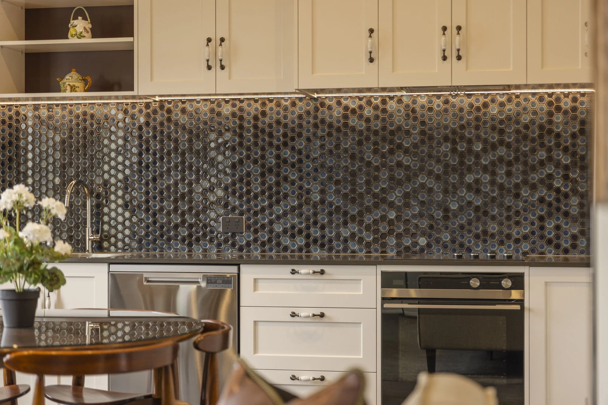 Kitchen with white cabinets, a mosaic backsplash with hexagon tiles, and stainless steel appliances, including a dishwasher and oven. There are decorative teapots on open shelves and a small table with chairs in the foreground.