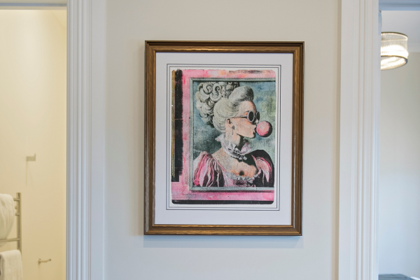 Framed artwork of a woman with curly hair, wearing sunglasses and a pink shirt, blowing a pink bubble gum, hanging on a white wall in a room.