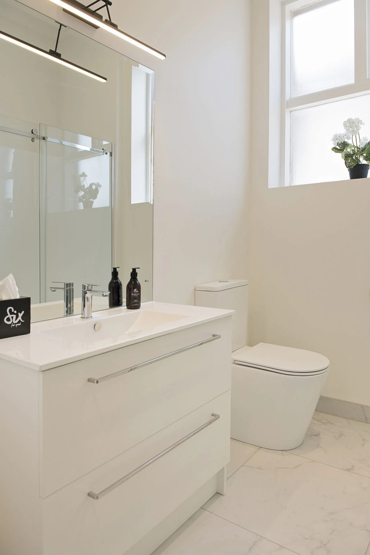 A modern, minimalistic bathroom with white walls, a white vanity with two drawers, a large mirror, a glass-enclosed shower, a white toilet, a tall window with natural light, and a potted plant on the windowsill.