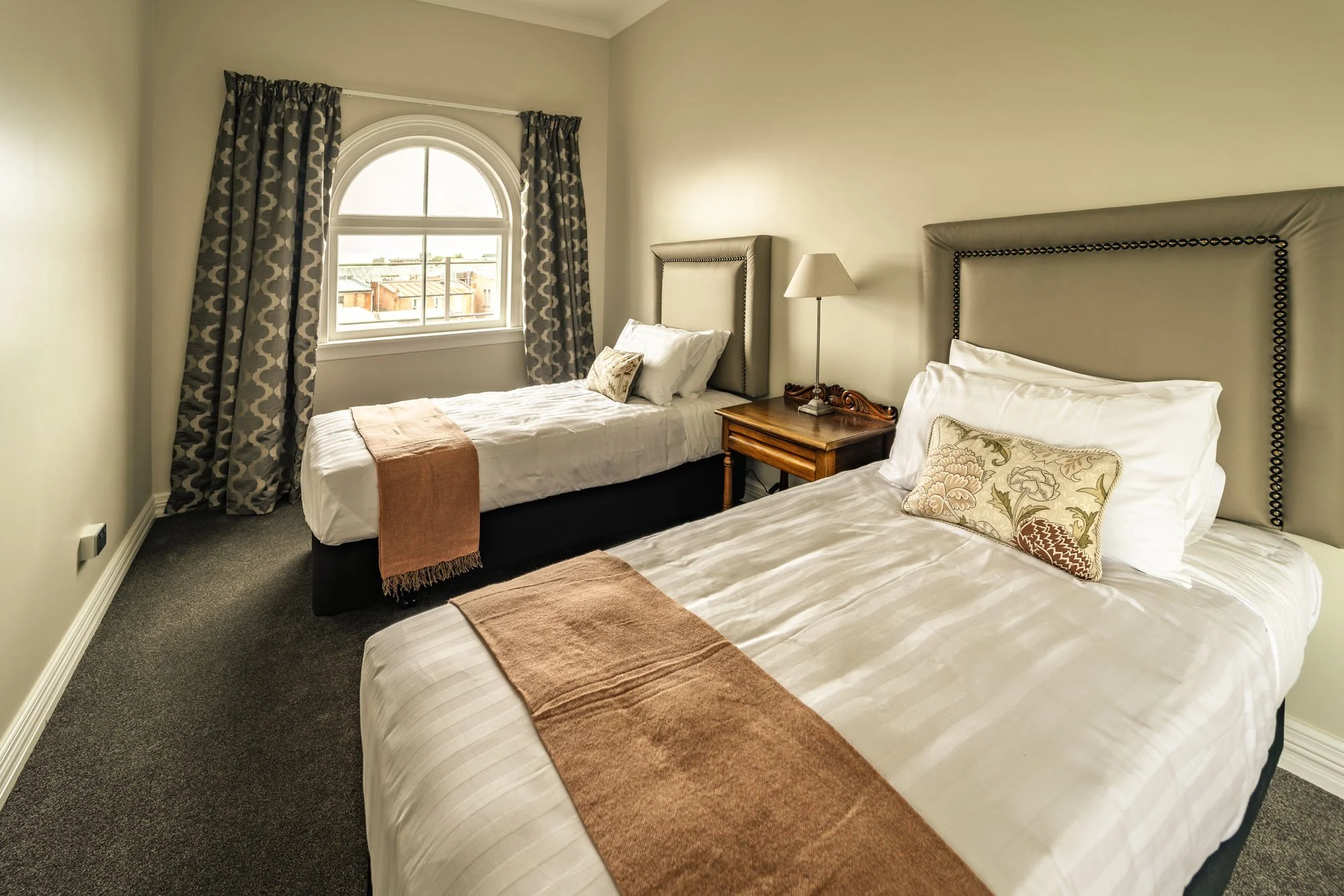 A hotel room with two beds, a window with patterned curtains, a nightstand with a lamp, and decorative pillows.