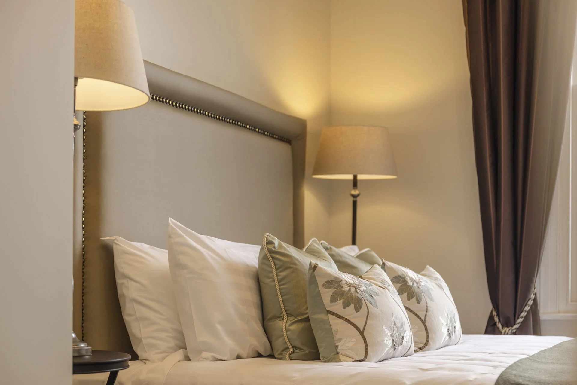 A neatly made bed with white sheets and multiple decorative pillows, flanked by two lamps and dark curtains, in a cozy bedroom setting.