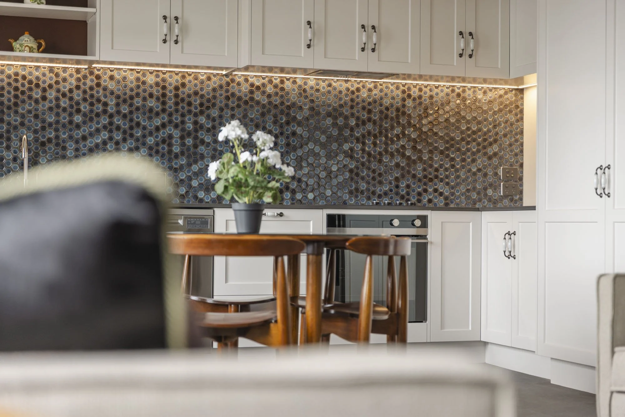 Modern kitchen with white cabinets, wooden dining table with chairs, black potted plant, honeycomb-patterned backsplash, and gray appliances.