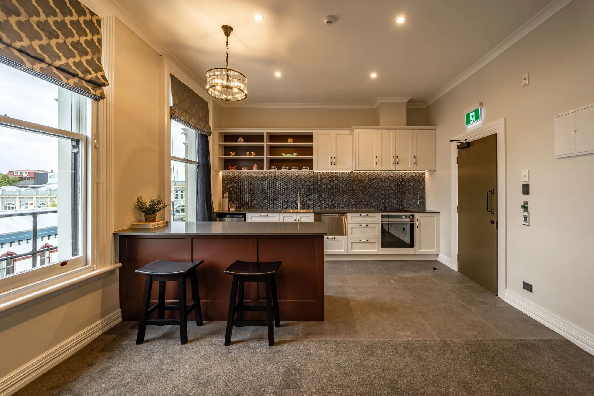 Modern kitchen with white cabinets, a dark mosaic backsplash, and a central island with two black stools. Large windows with patterned blinds, and a chandelier hanging from the ceiling.