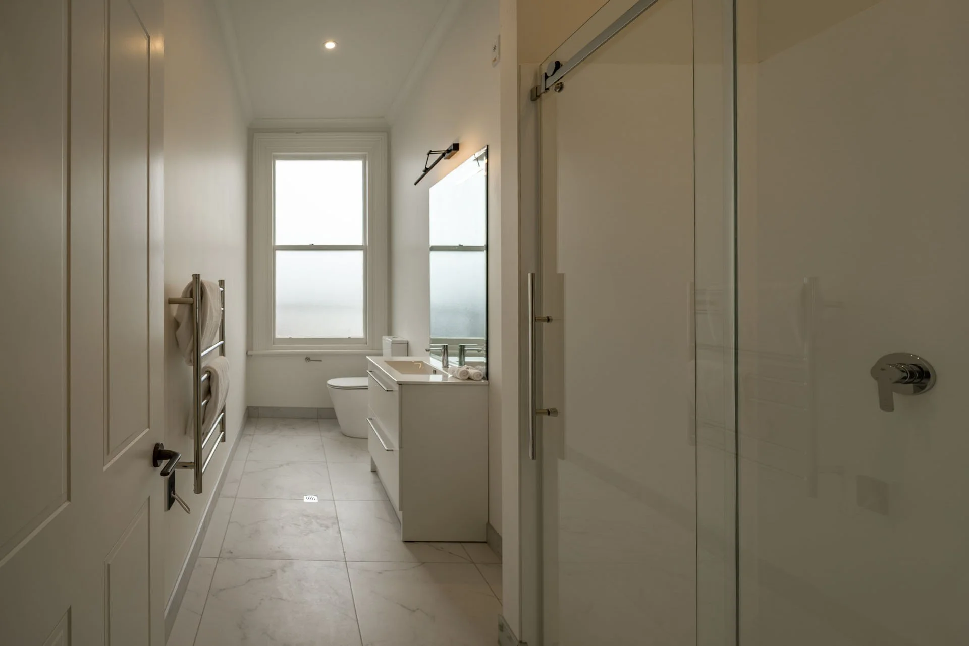 Modern bathroom with white walls, tiled floor, window, toilet, vanity with sink, mirror, towel rack, and glass shower enclosure.