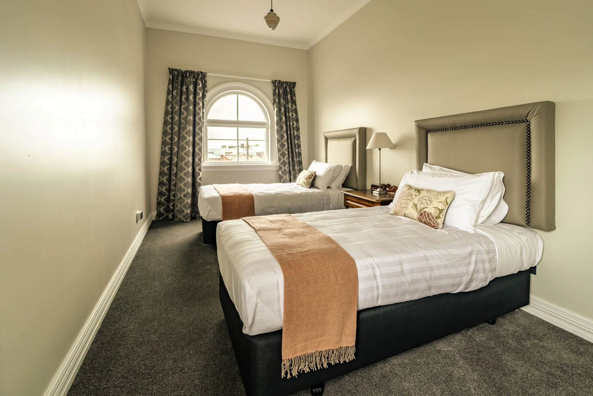 A hotel room with two beds, each with beige headboards, white bedding, beige and patterned decorative pillows, tan throws on top, a bedside table with a lamp, and a window with patterned curtains.