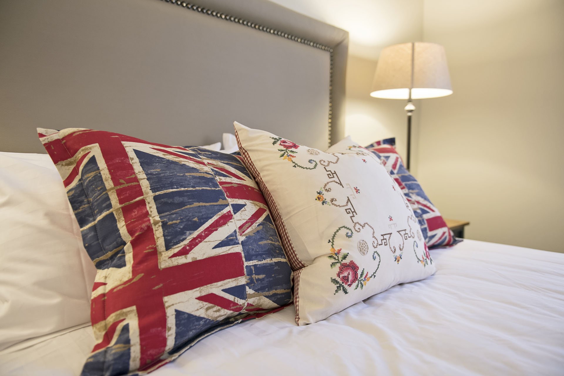 Decorative pillows on a bed, with one featuring a Union Jack flag design, another with embroidered flowers and patterns, and a third with additional Union Jack pattern, next to a bedside lamp.
