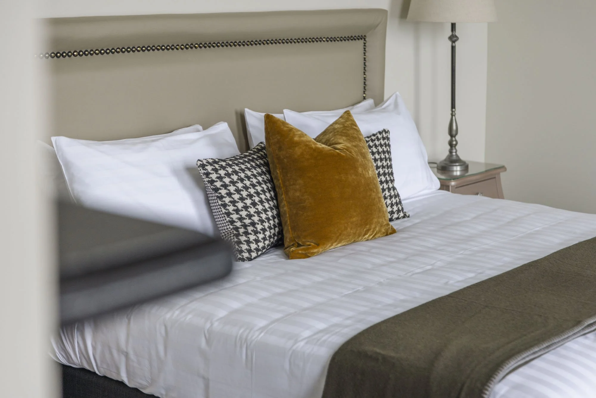 A neatly made bed with white sheets, a brown blanket, and decorative pillows including a brown plush pillow and black-and-white houndstooth patterned pillows, with a beige headboard and a bedside lamp on a nightstand.