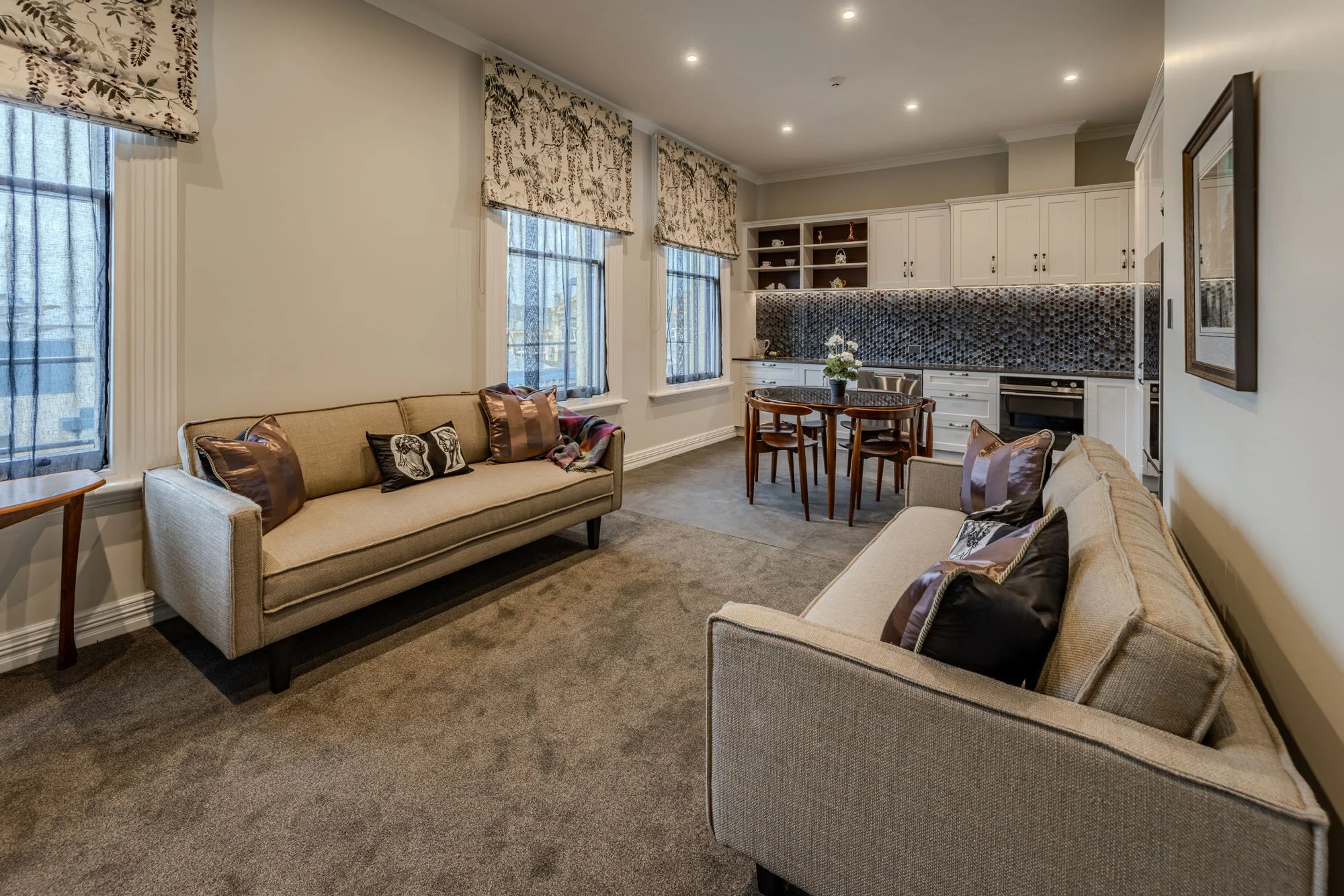 Living room with beige sofas, patterned curtains, and a kitchen area with white cabinets, a round dining table, and black backsplash tiles.