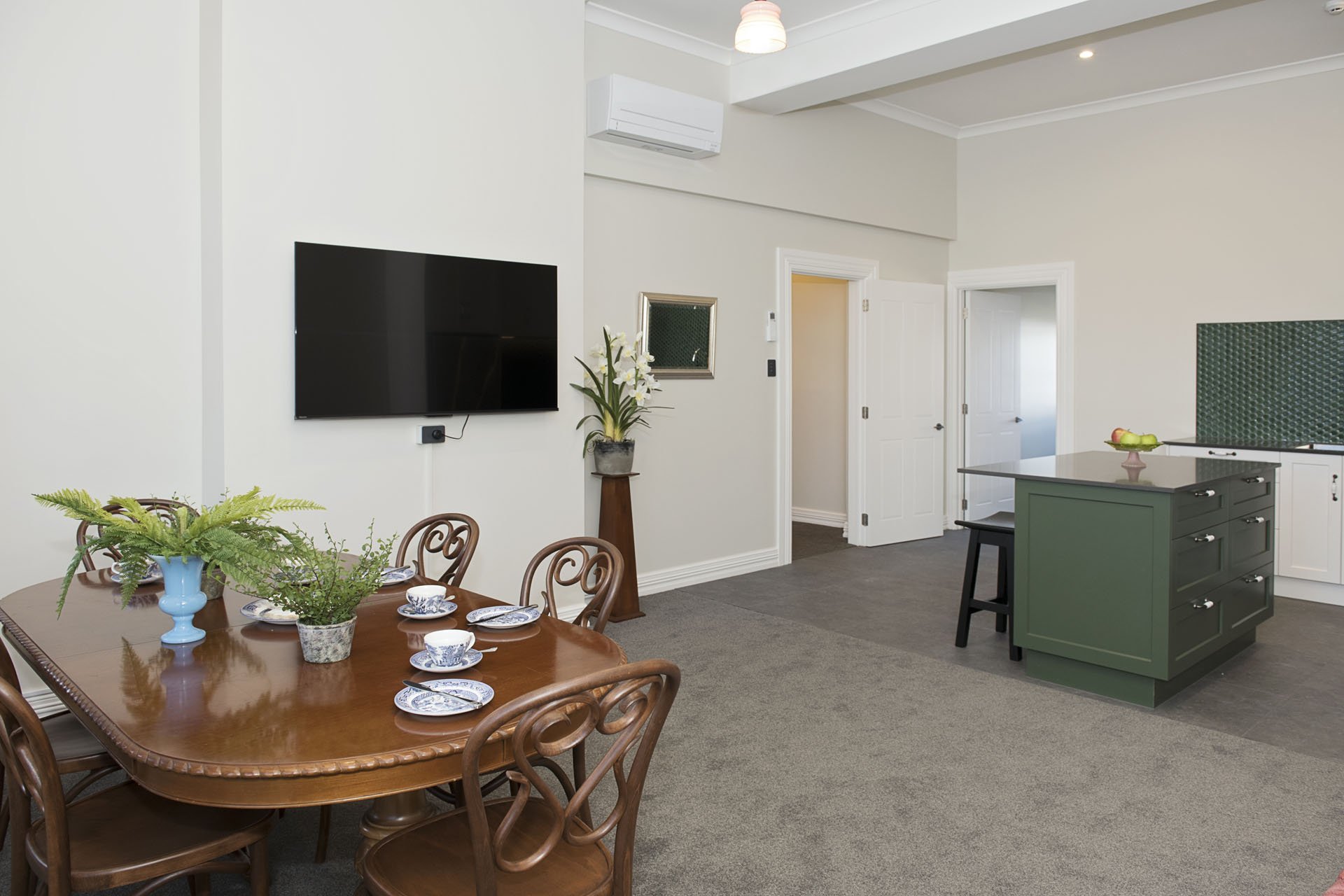 A dining area with a wooden dining table set for four, decorated with houseplants in ceramic and glass pots, in a room with a mounted television, white walls, and neutral flooring, adjacent to a kitchen island with green cabinetry.