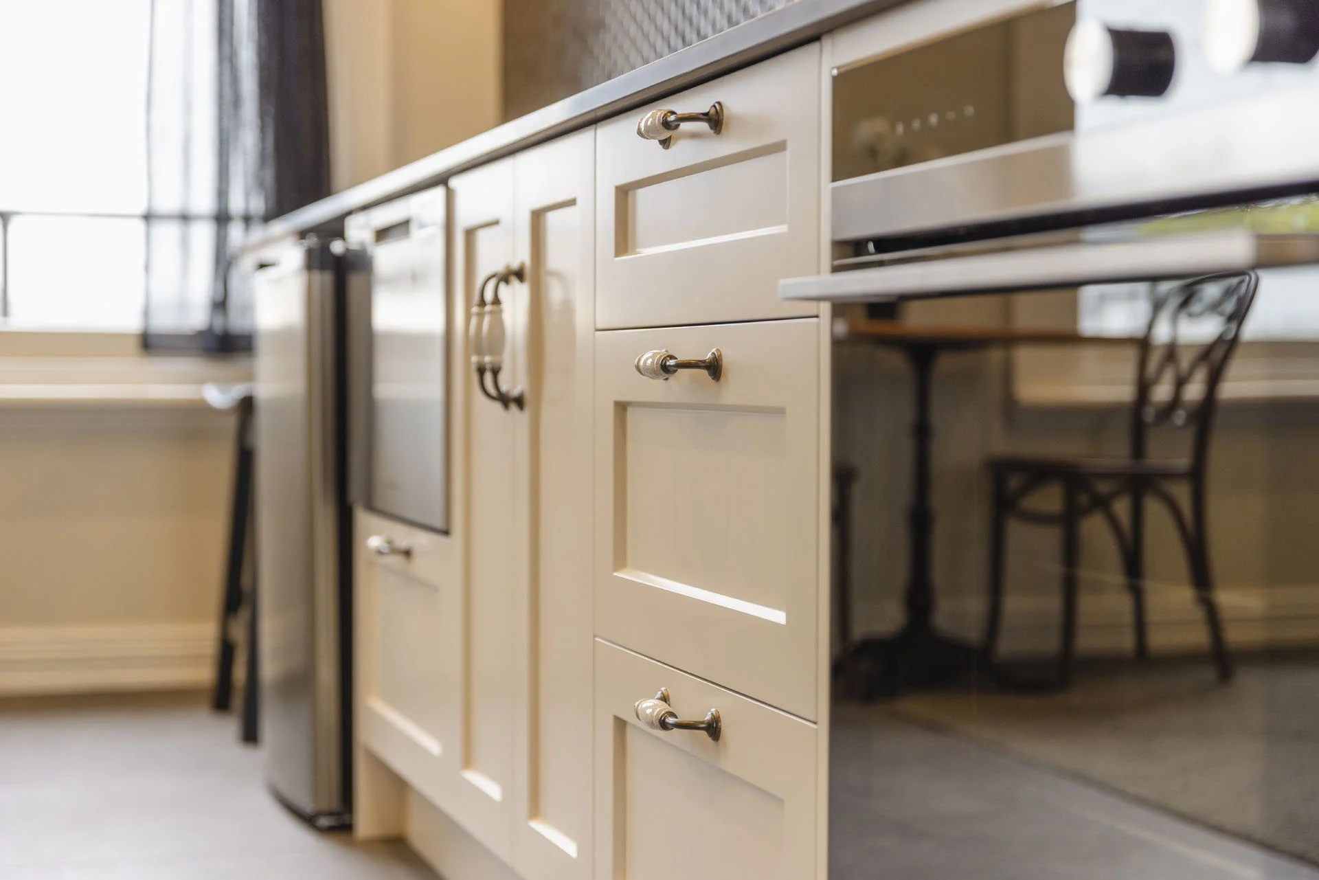 Close-up of a white kitchen cabinet with silver handles, a black chair reflection in the oven window, and a window with a grid inside a modern kitchen.