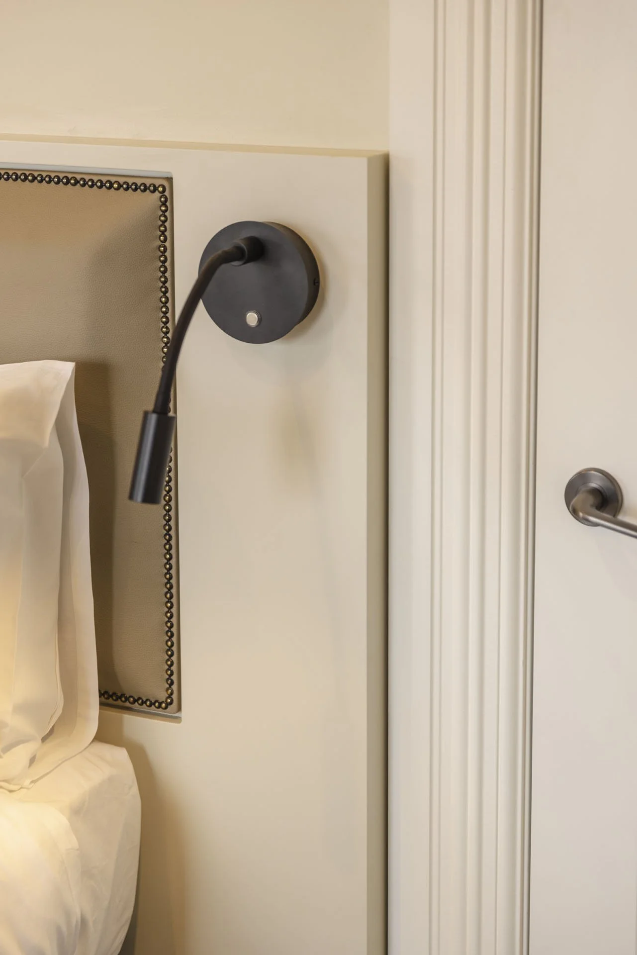 Close-up of a bedroom nightstand with a wall-mounted reading lamp and part of a bed with white bedding.