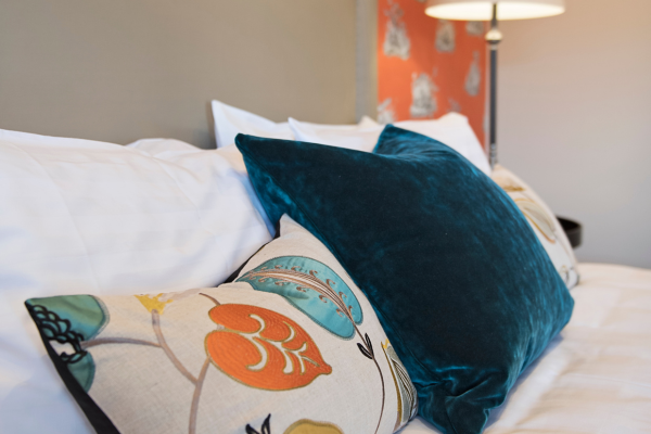Close-up of a bed with white pillows, a dark teal velvet pillow, and a decorative pillow with a floral pattern. A lamp and a patterned accent wall are in the background.