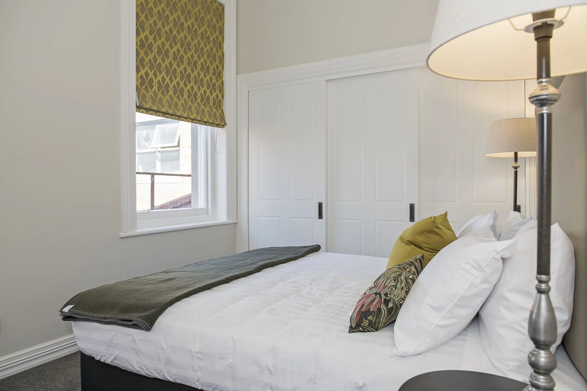 A neatly made bed in a bedroom with white walls, a window with a geometric-patterned yellow blind, two lamps on nightstands, and a closet with sliding doors.