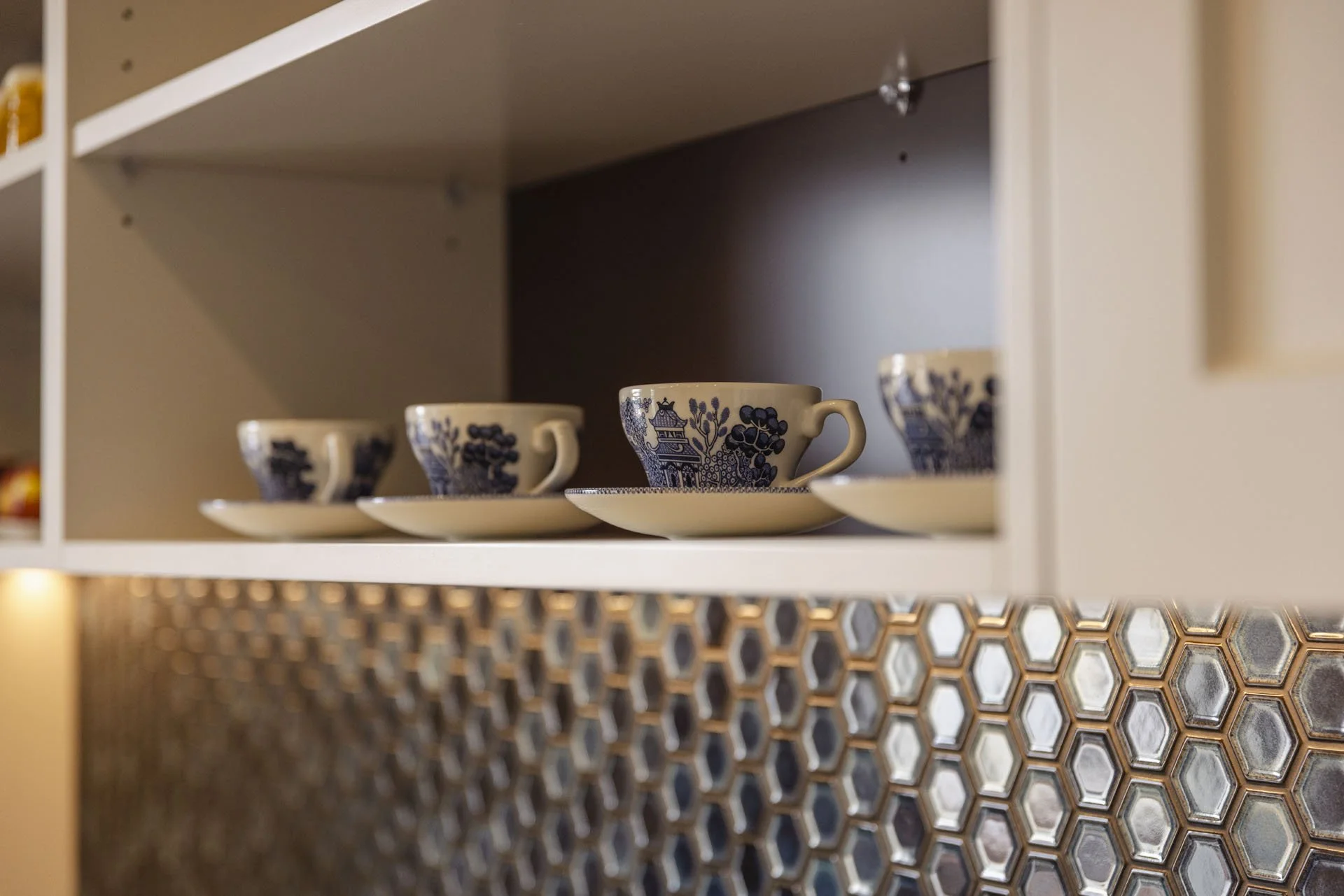 Three teacups with matching saucers on a kitchen shelf, decorated with blue floral and building patterns, and a honeycomb-patterned backsplash below.