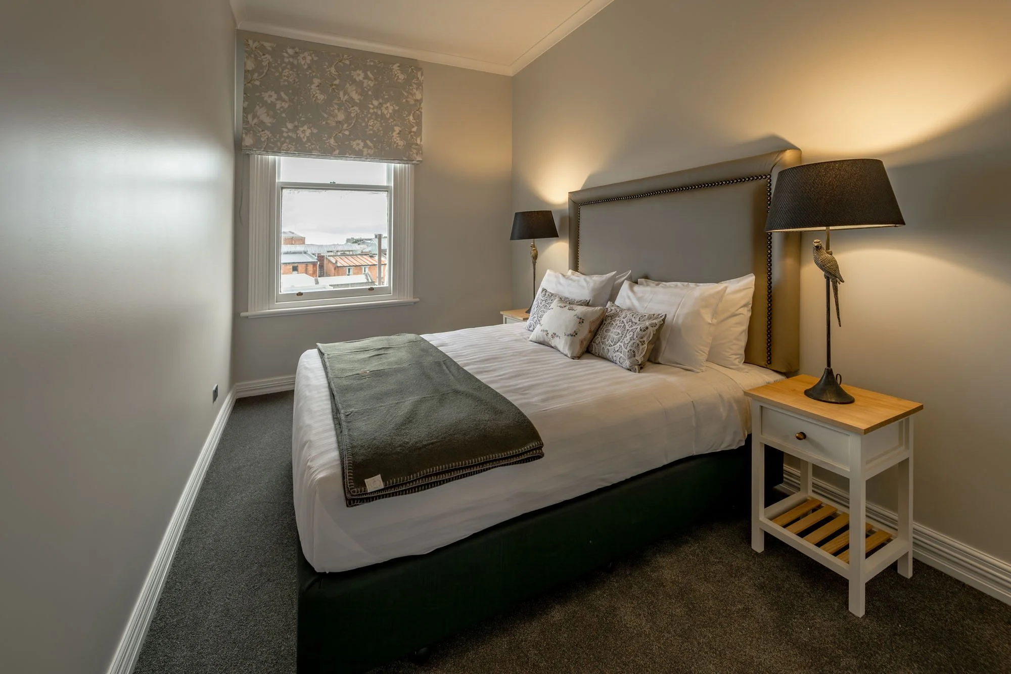 A cozy bedroom with a large bed, decorative pillows, nightstands with lamps, and a window with a view of rooftops.