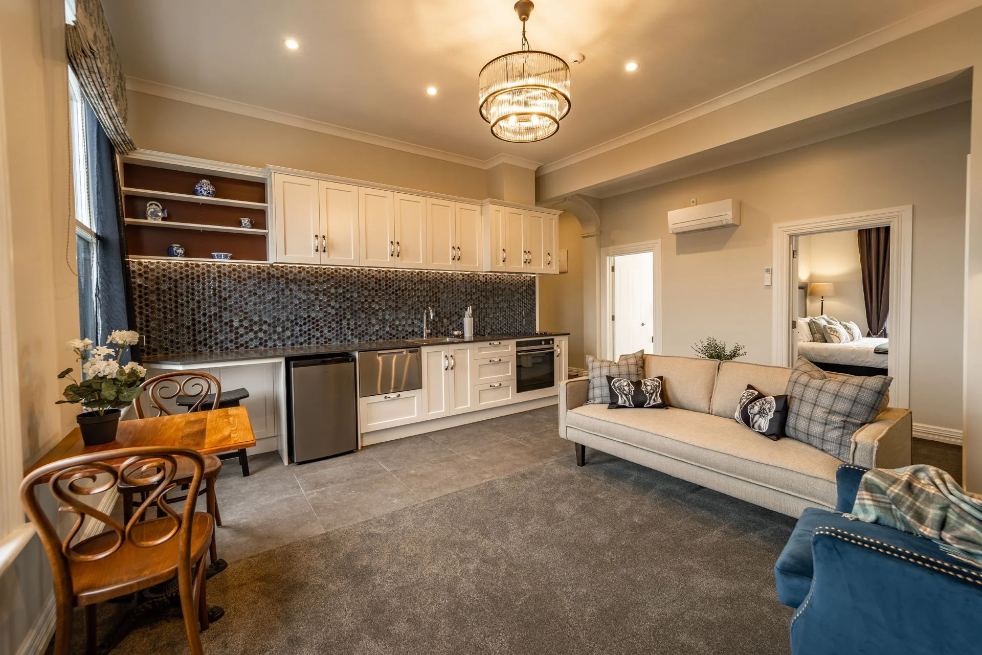 Open-concept living room and kitchen with beige walls, white cabinets, and dark mosaic tile backsplash. There is a small dining table with three wooden chairs, a beige sofa, a blue armchair, and a bedroom visible through a doorway. A chandelier hangs