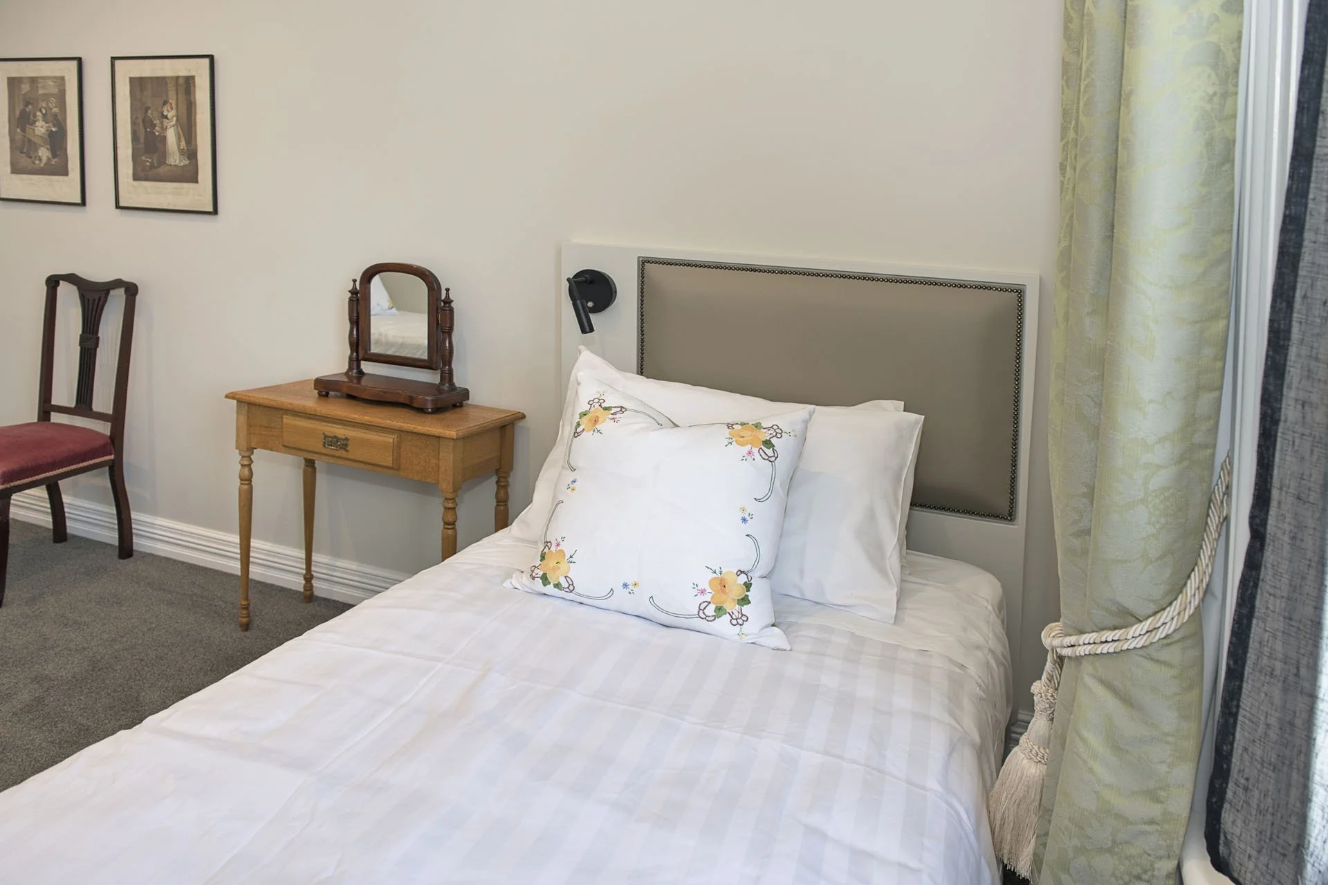 A neatly made bed with a embroidered pillow and beige headboard, next to a window with yellow curtains. To the left, a small wooden table with a vintage mirror and a wooden chair with a red cushion, hanging picture frames on the wall.