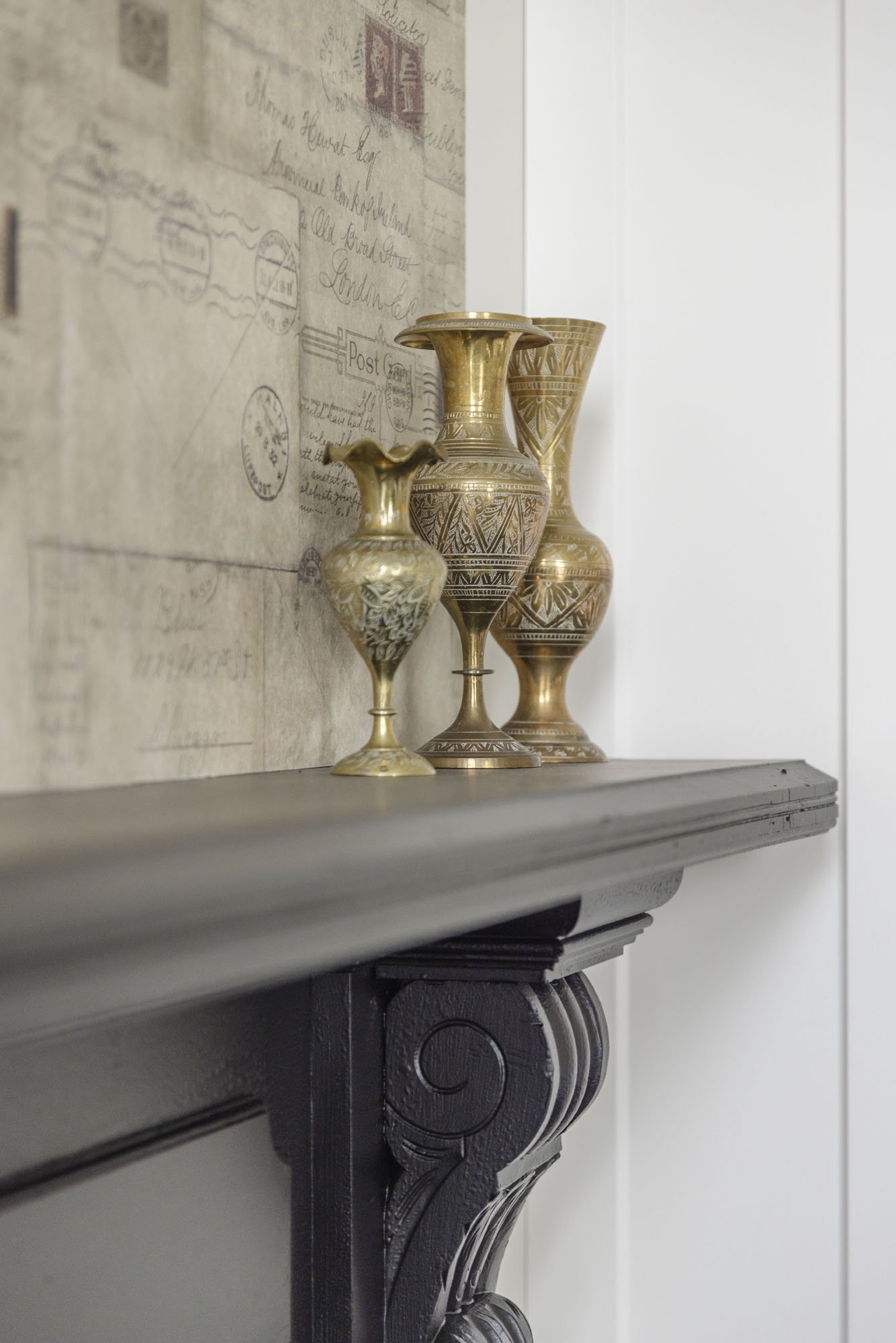 Three gold and brass decorative vases placed on a dark fireplace mantel against a wall with vintage postcard and letter prints.