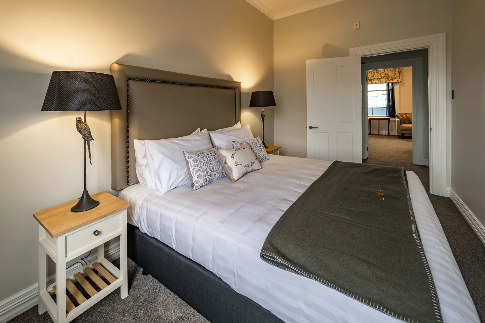 A cozy hotel bedroom with a large bed, decorative pillows, two lamps with bird-shaped bases, and an open door revealing a sitting area with a couch and window.