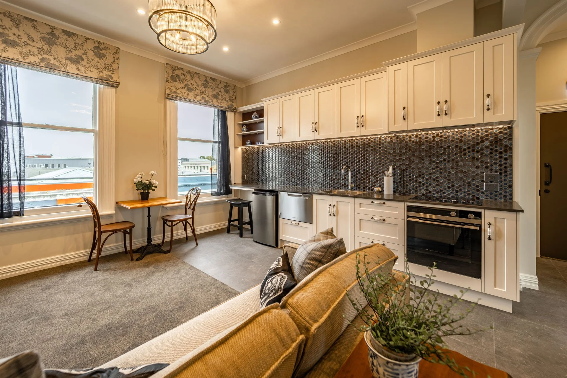 Interior view of a living space with a cozy sofa, a kitchen with white cabinets, dark backsplash, and modern appliances, two large windows with blue curtains, a small wooden table with two chairs by the window, and decorative plants.
