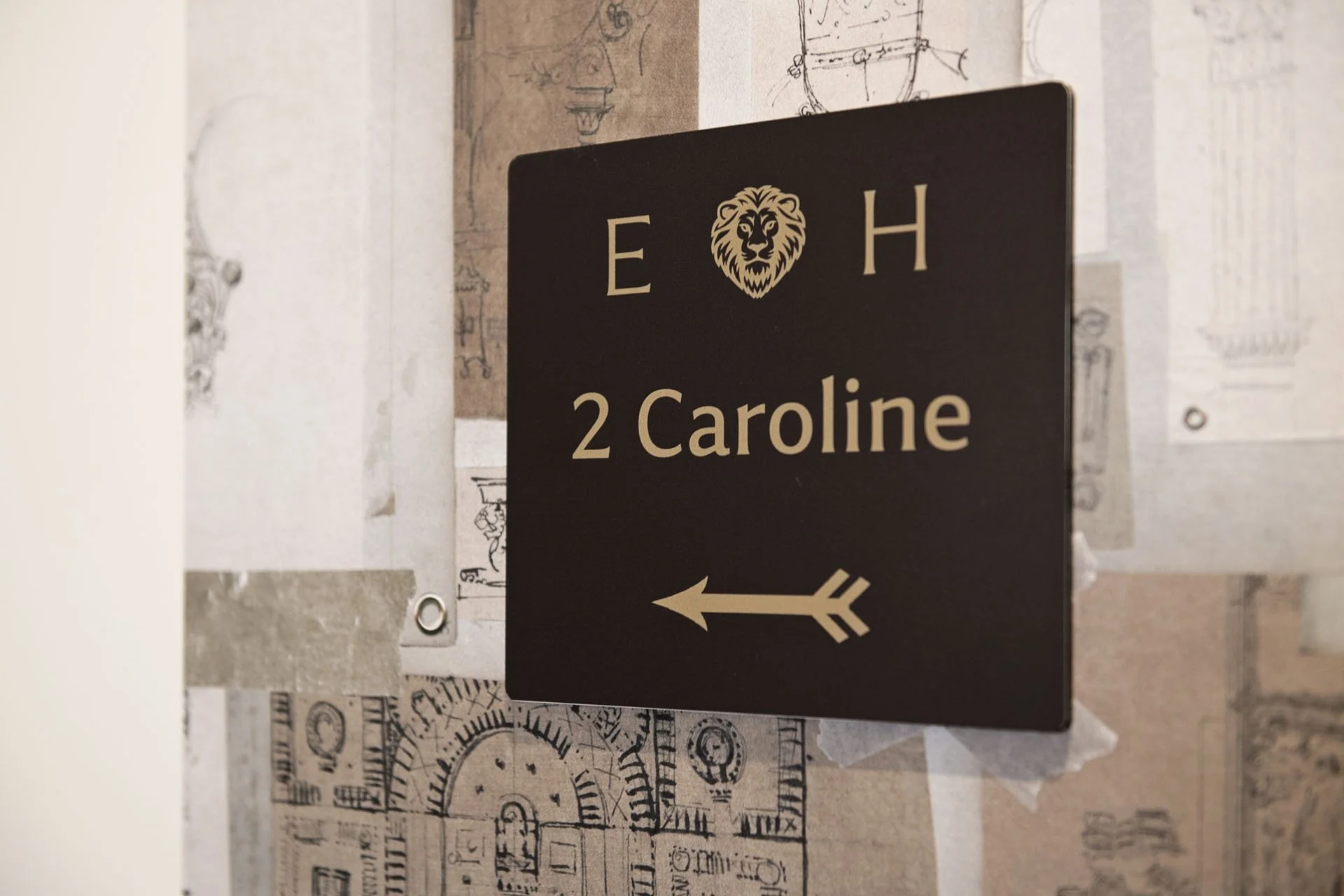A black sign with a lion's head in the center above the text '2 Caroline' and an arrow pointing to the left, mounted on a wall with doodles and sketches.