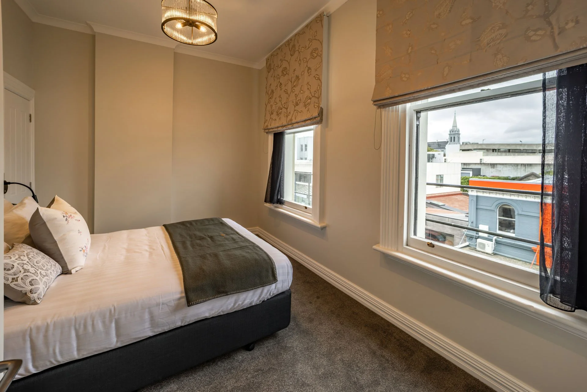 A bedroom with a bed, two windows with curtains, a view of buildings and a church steeple outside.