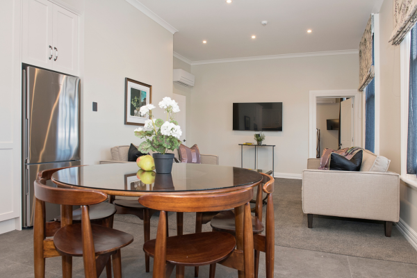 Living and dining area with a round wooden table, four chairs, a floral centerpiece, a beige sofa, a wall-mounted TV, and a small side table with a plant.