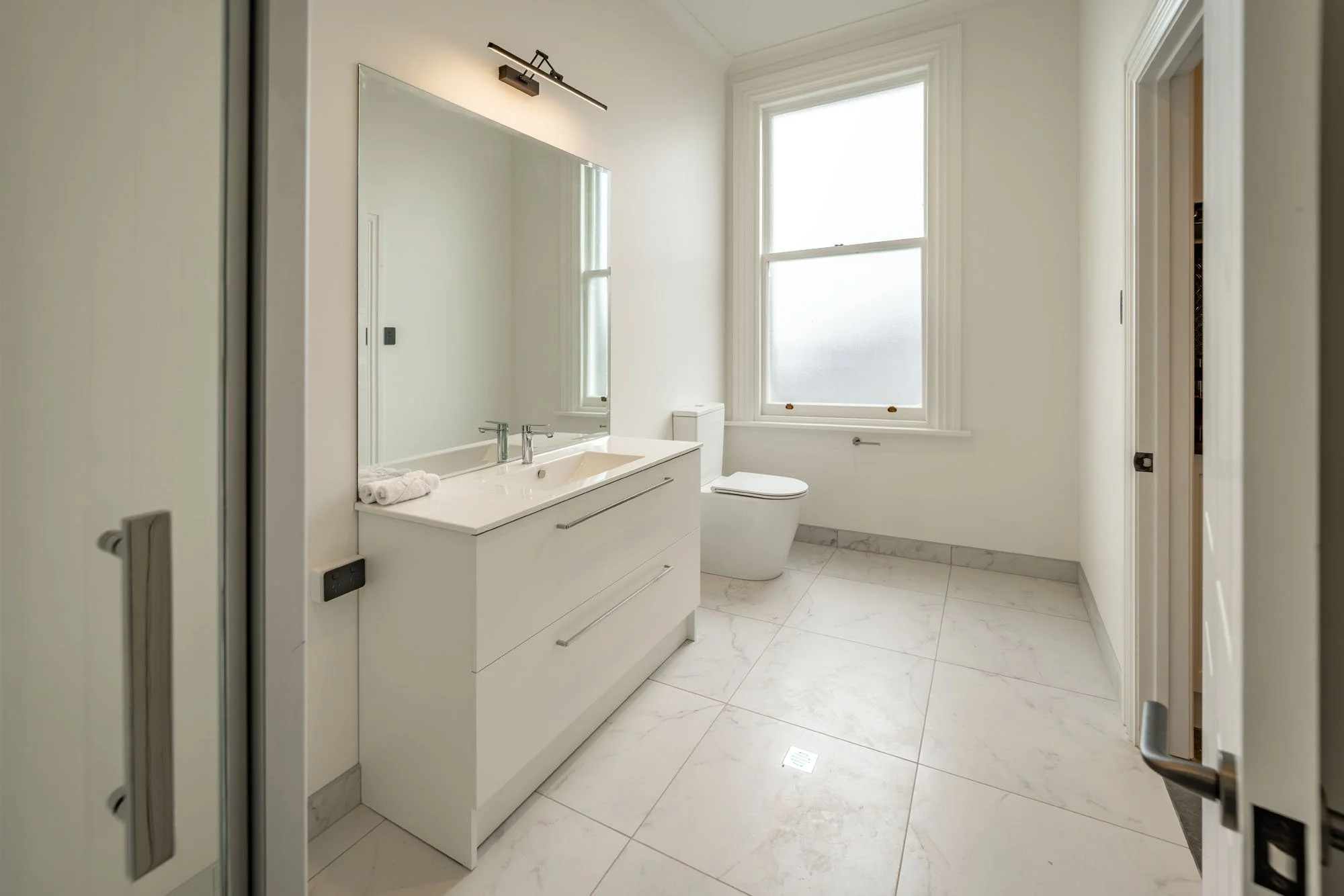A clean, modern bathroom with white walls, a large mirror, a double sink vanity, a toilet, and large windows letting in natural light, with marble flooring.