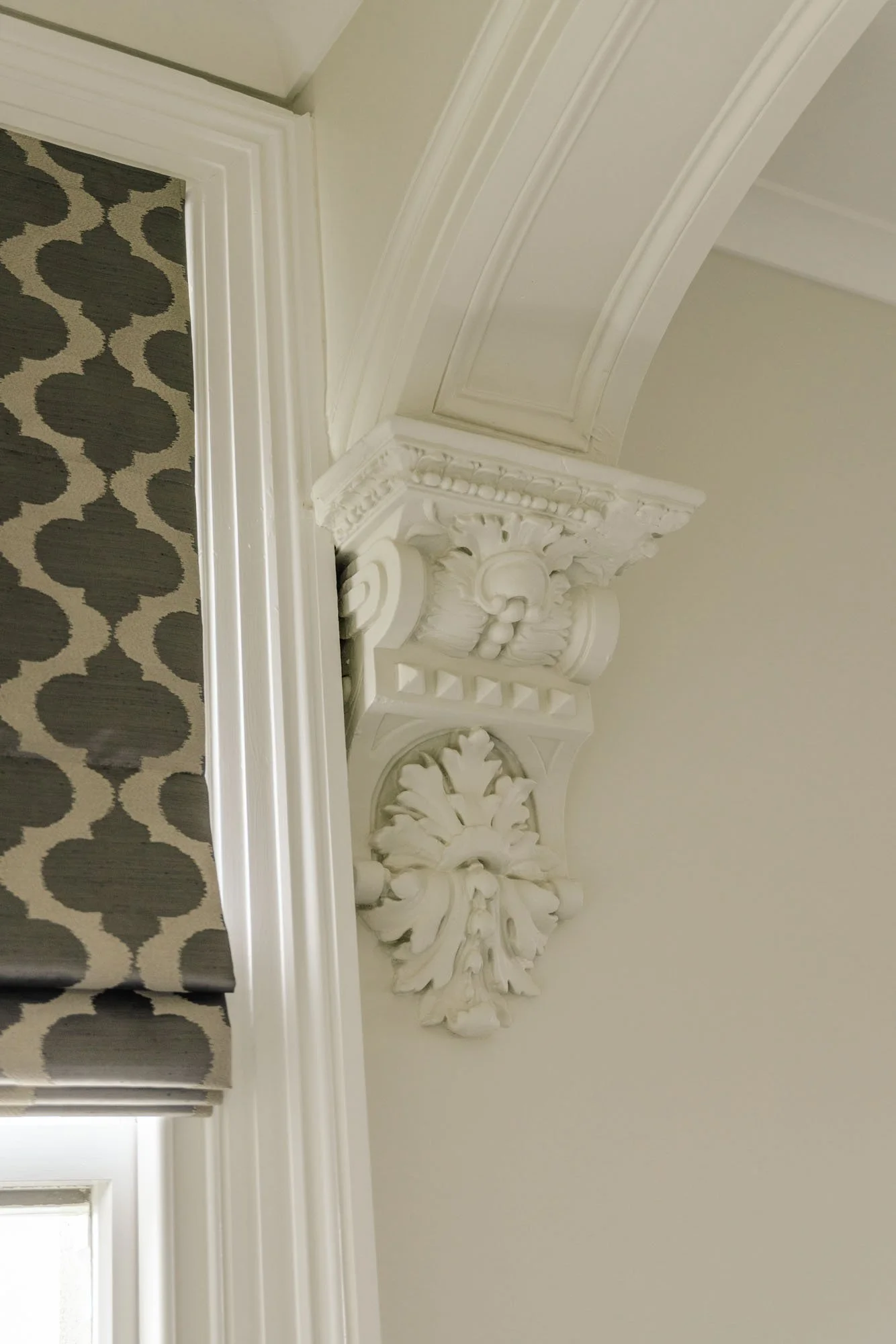 Close-up of ornate white decorative crown molding and wall corner next to window with patterned curtains.