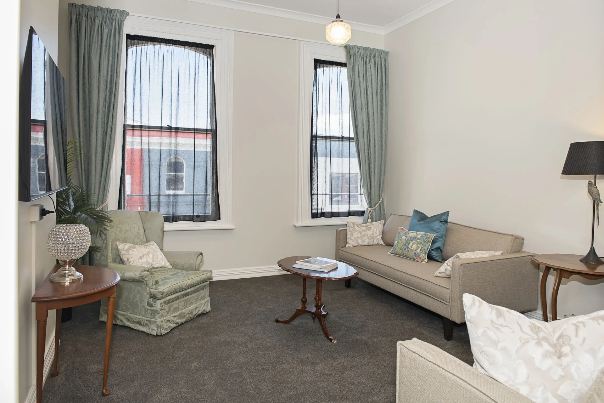 Living room with beige sofa, an armchair with green floral fabric, a small wooden coffee table, side tables, a wall-mounted TV, windows with black lace curtains, and blue and white decorative pillows.