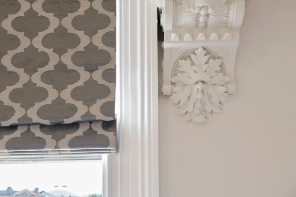 Close-up view of an ornate white crown molding and decorative architectural detail on a wall, next to a window with a patterned gray Roman shade.