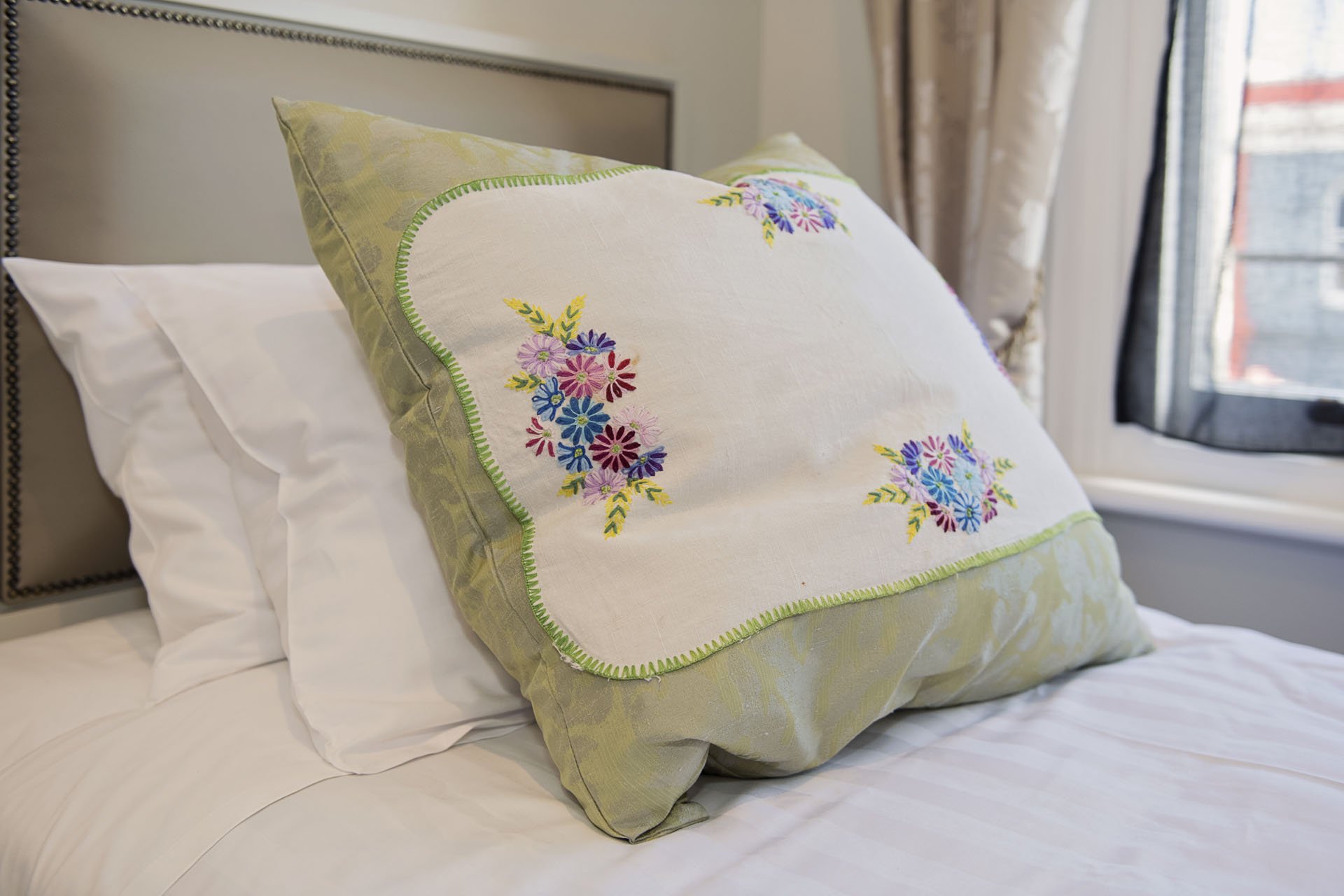 Decorative embroidered pillow with floral pattern on a bed with white sheets, near a window with curtains.