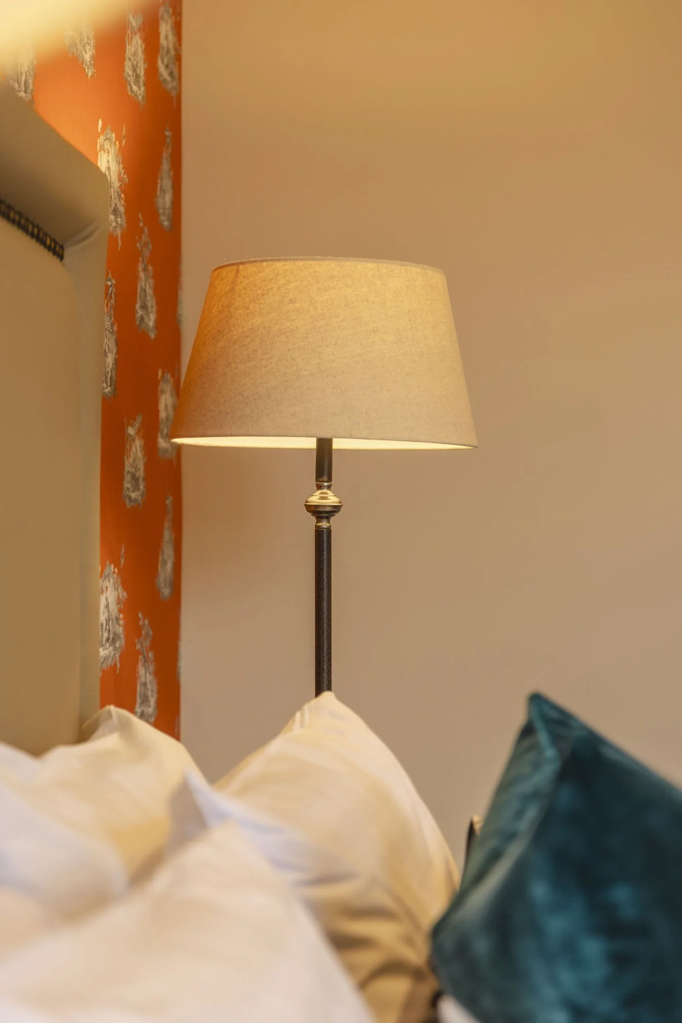 A bedroom corner with a bedside lamp on a beige wall, cream-colored pillows, and a teal decorative pillow in the foreground. An orange textured wall with gray floral patterns is visible in the background.