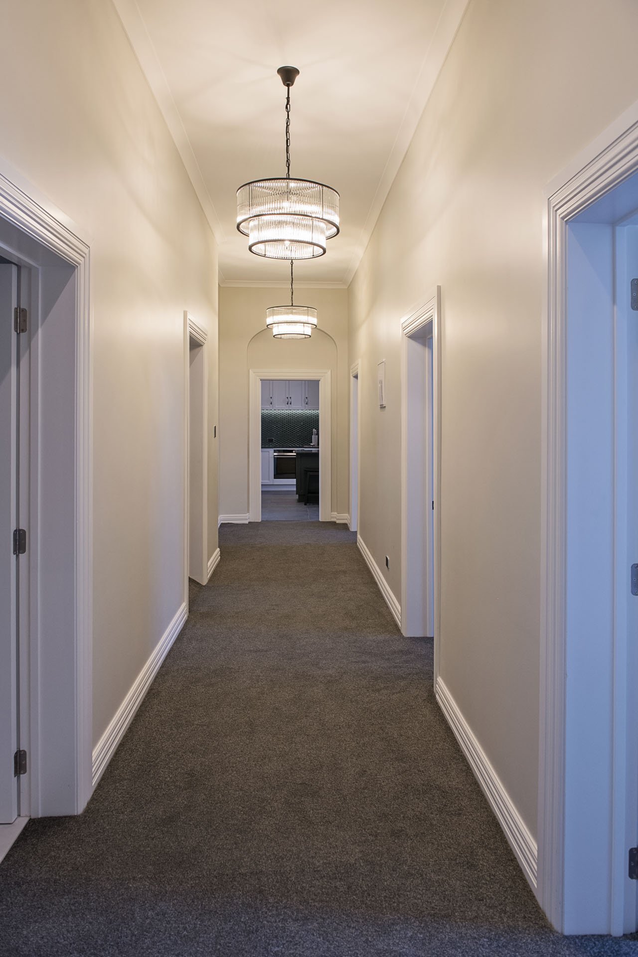 A hallway with beige walls, white crown molding, and a brown carpet runner. There are four modern ceiling lights hanging from the ceiling. At the end of the hallway, an open doorway leads to a kitchen area.
