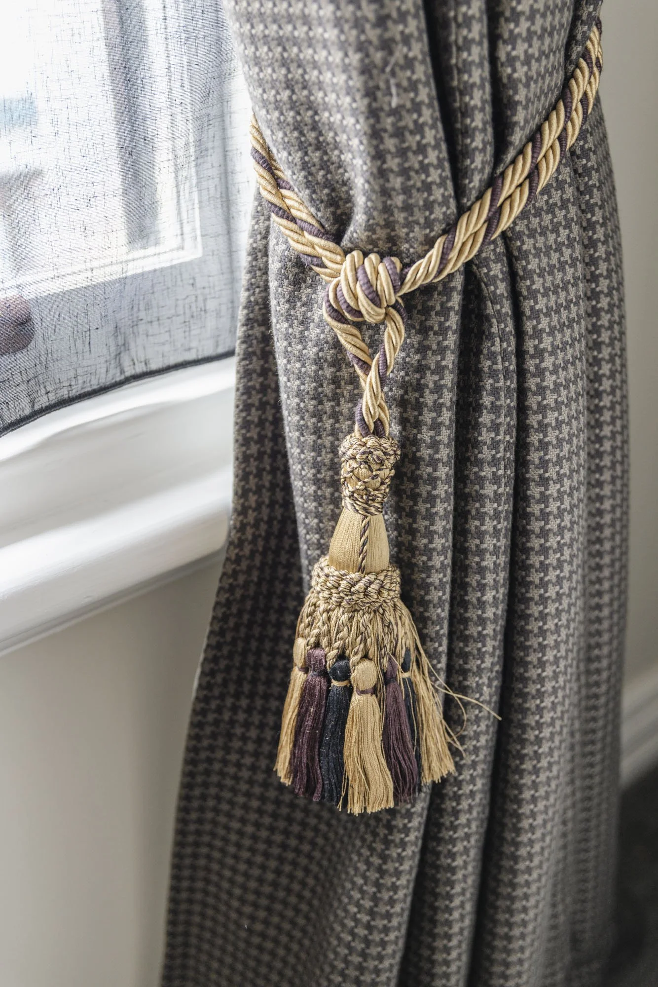 Decorative tassel tie-back on a patterned gray curtain near a window.