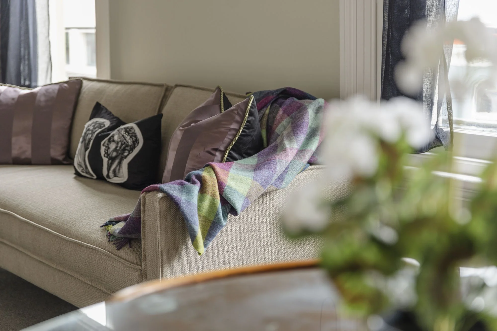 Living room with a beige sofa adorned with various pillows, a blanket draped over the armrest, and a window with dark curtains in the background.