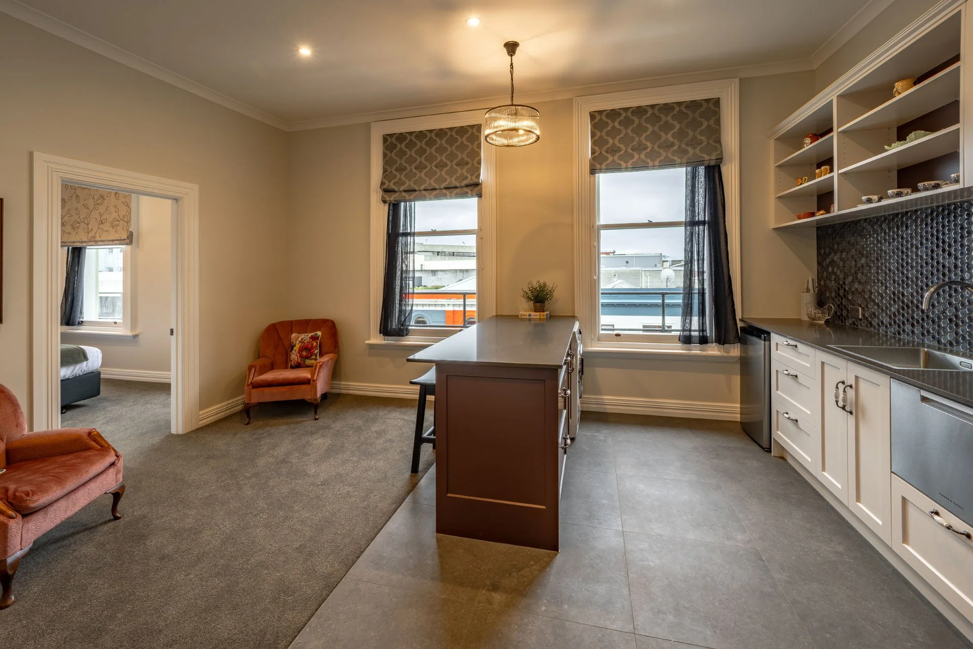 Kitchen and living room with two vintage pink armchairs, large windows with patterned shades, a kitchen island, white cabinets, open shelving with dishes, gray tile backsplash, and gray flooring