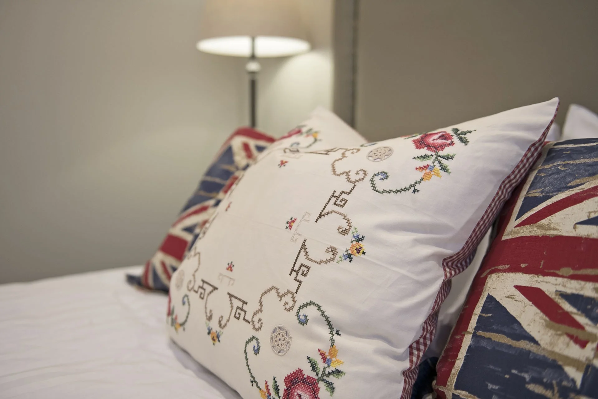 Decorative pillows with Union Jack design and embroidered pillow with cross-stitched floral pattern and the words 'Peace, Love, Joy' on a bed next to a bedside lamp.