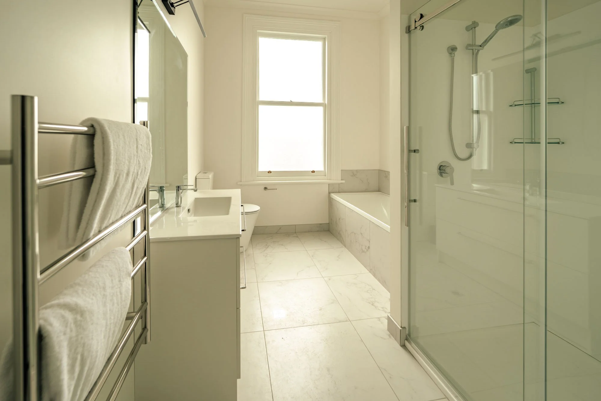 A bright bathroom with a large window, a bathtub, a glass shower enclosure, a white vanity with a sink, a toilet, and a heated towel rack with towels.