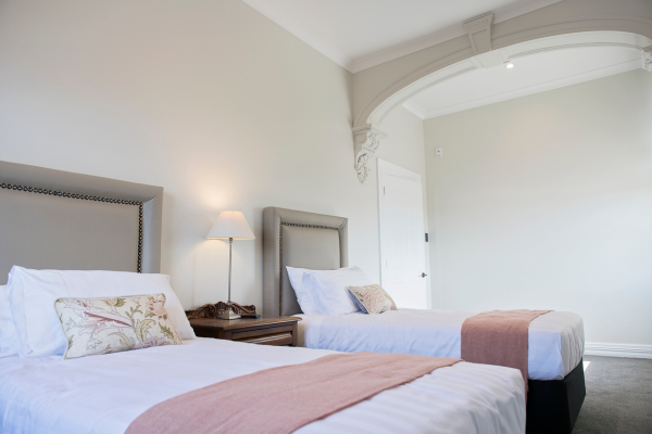 Two neatly made beds with white linens and decorative pillows in a bright hotel room, with a wooden nightstand, a lamp, and arched window or doorway.