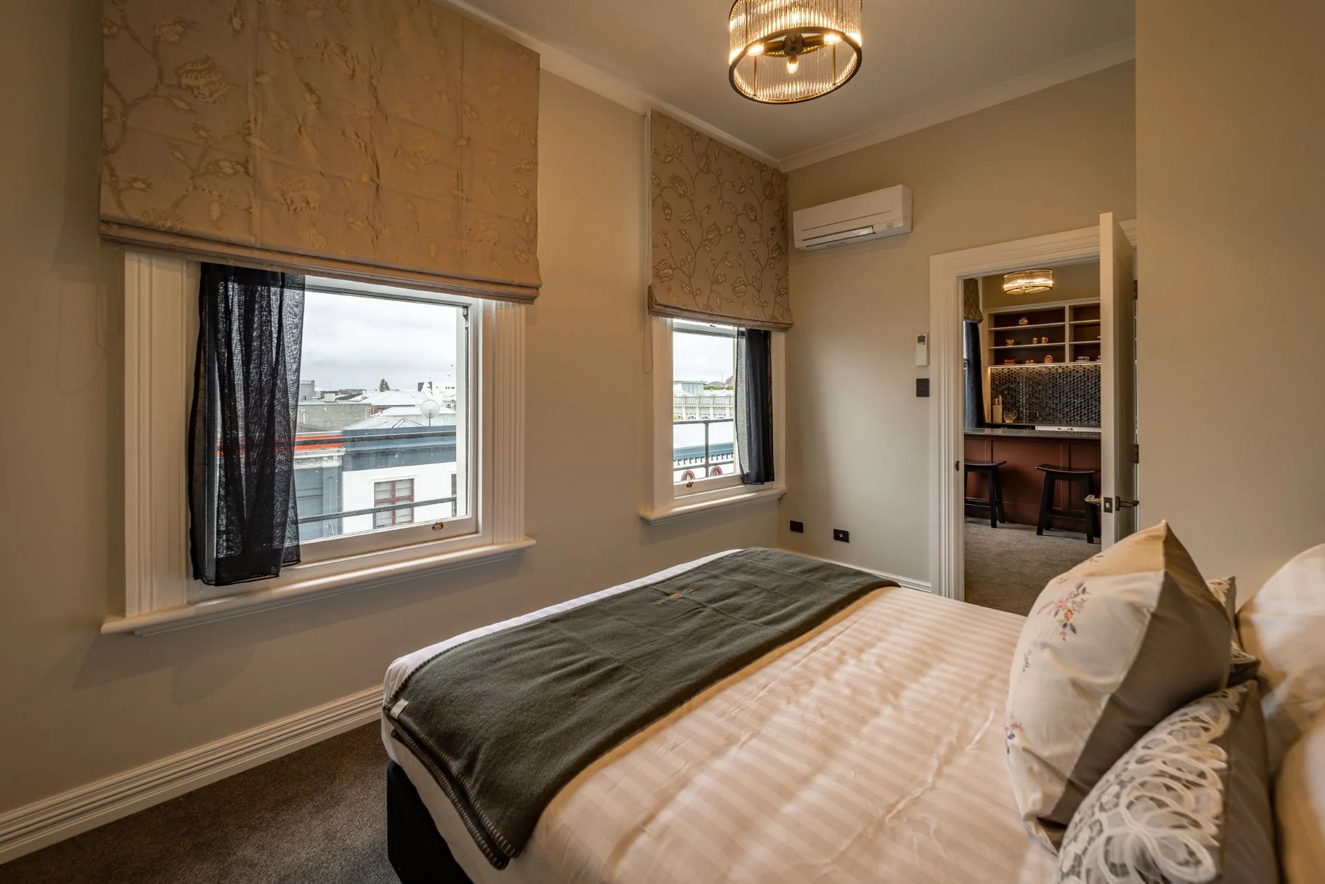 A cozy bedroom with two windows, beige walls, and a bed with striped bedding and pillows. There is a doorway leading to a small living area with a bar and two stools, and an air conditioning unit mounted on the wall.