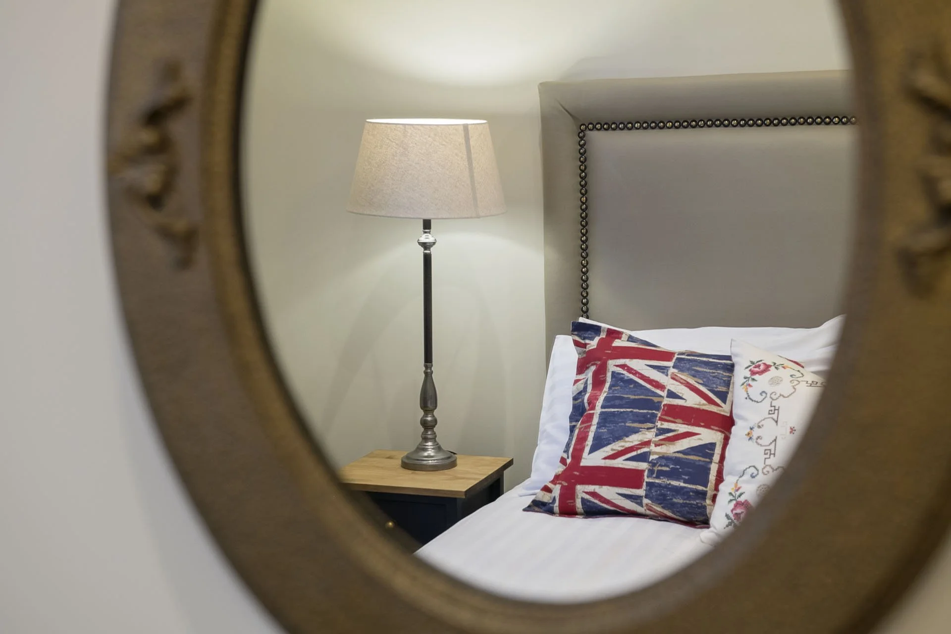Reflection in an ornate gold-framed mirror showing a bedroom corner with a bedside lamp, a headboard with studded trim, a pillow with a Union Jack print, and part of a wood nightstand.